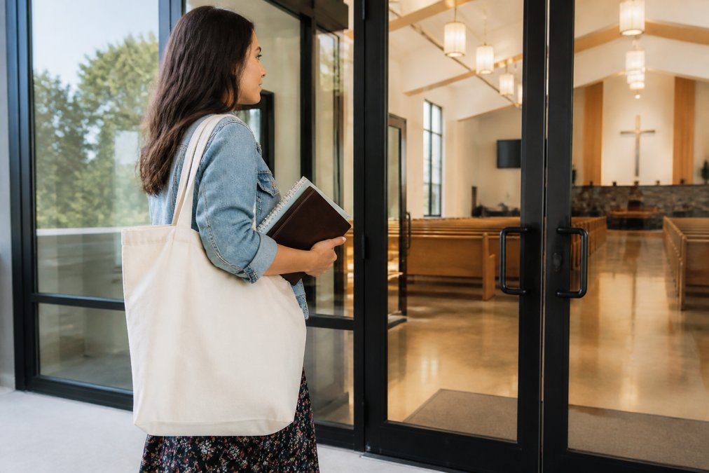 A woman carrying a natural canvas tote bag with a faith-themed design, walking into a bright modern church building, holding a Bible and a small notebook