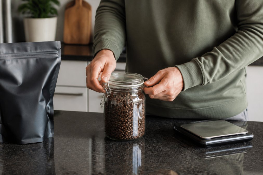 Person sealing an airtight jar of fresh roasted coffee beans on a kitchen counter with a digital scale nearby