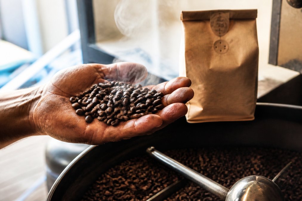 Close-up of a coffee roaster's weathered hand holding a handful of freshly roasted dark brown beans with visible steam rising, with a kraft paper bag displaying a printed roast date visible in the soft background light