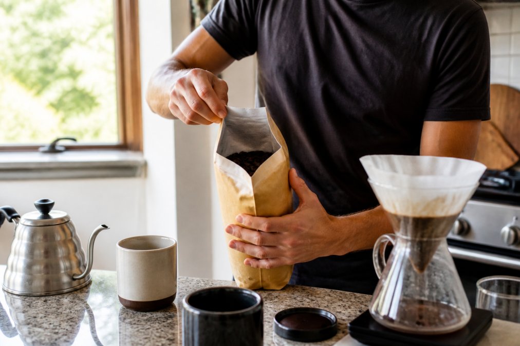 A person opening a delivered coffee bag at a kitchen counter, whole beans visible, pour-over setup nearby, soft morning light through the window