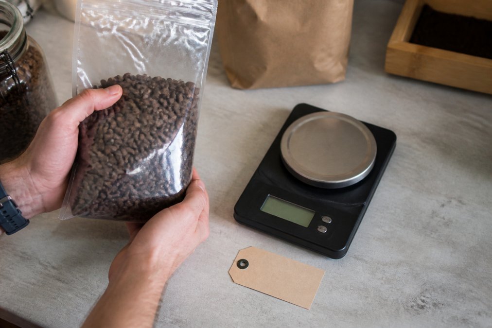 Overhead shot of a person's hands holding a clear bag of whole bean coffee next to a digital scale and a printed roast date label on a kitchen counter