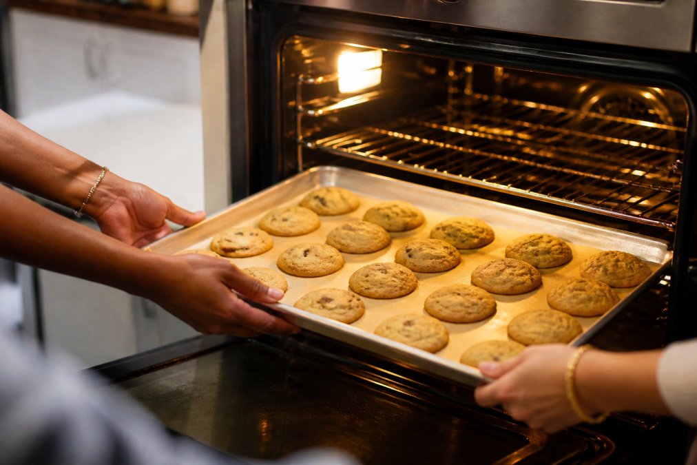 Close-up of hands showing different skin tones carefully placing a tray of golden-brown cookies into a warm oven, with kitchen light highlighting the texture and color of the baked goods