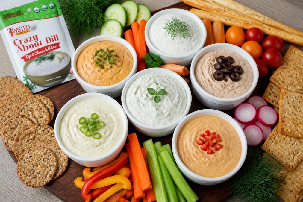 Overhead shot of a party platter with multiple colorful dips in white ceramic bowls, fresh vegetables, whole grain crackers, and breadsticks arranged on a wooden serving board with fresh herbs as garnish