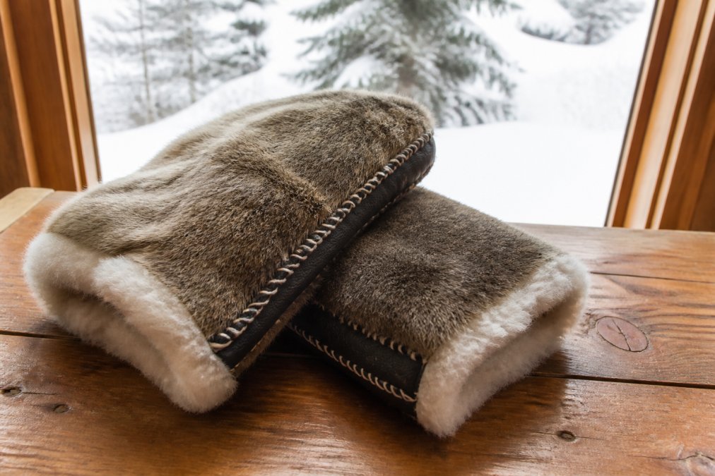 Close-up of a pair of handcrafted fur mittens with visible hand-stitching and a sheepskin lining, resting on a wooden table with snow visible through a window in the background