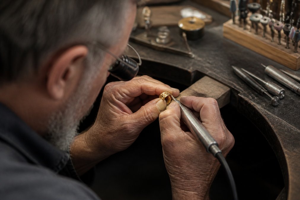Master jeweler's hands working on a delicate gold ring at a polished workbench, using precision tools and a jeweler's loupe, with soft studio lighting highlighting the intricate metalwork