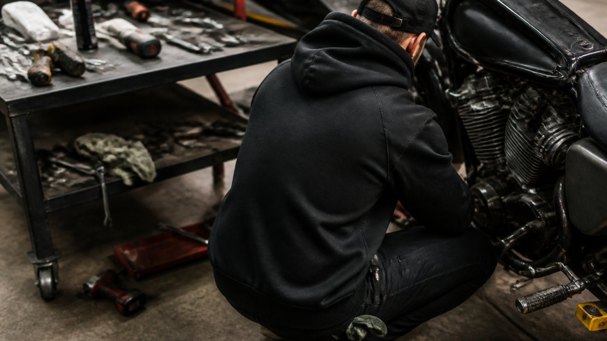 A mechanic wearing a black hoodie with a large back print, crouching beside a motorcycle in a garage workshop, tools scattered on a workbench nearby
