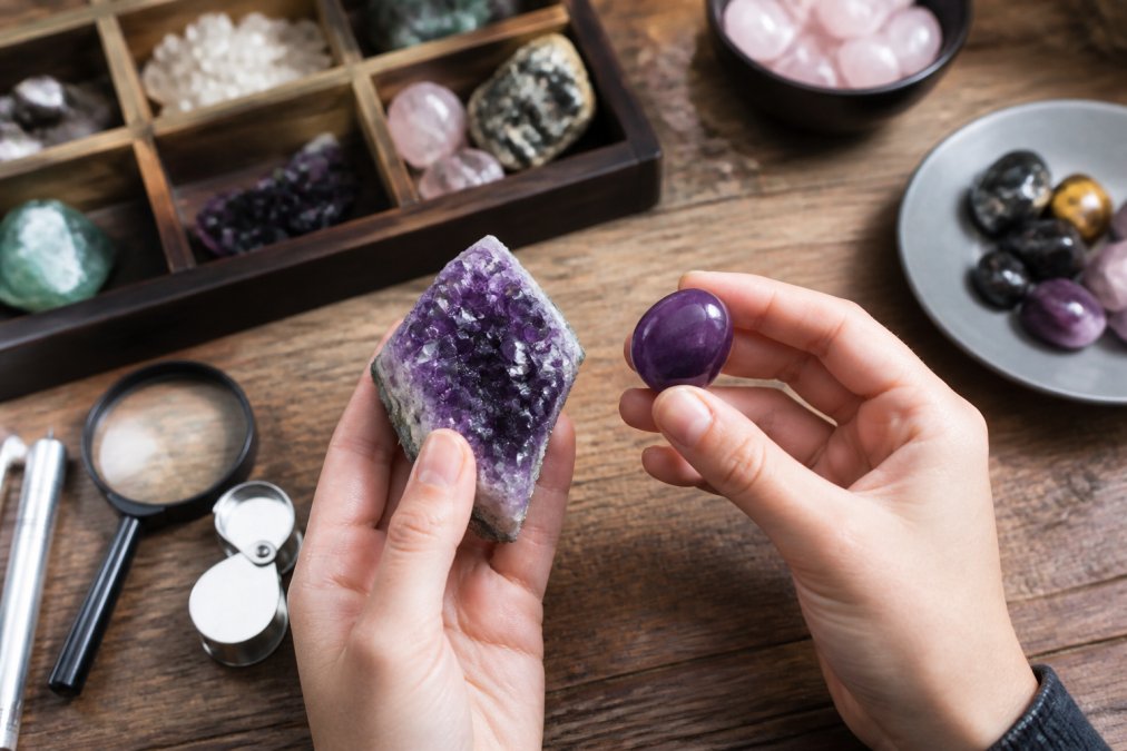 Close-up of two hands holding a raw amethyst geode and a polished tumbled stone side by side on a wooden surface, with soft natural lighting highlighting the texture and color differences
