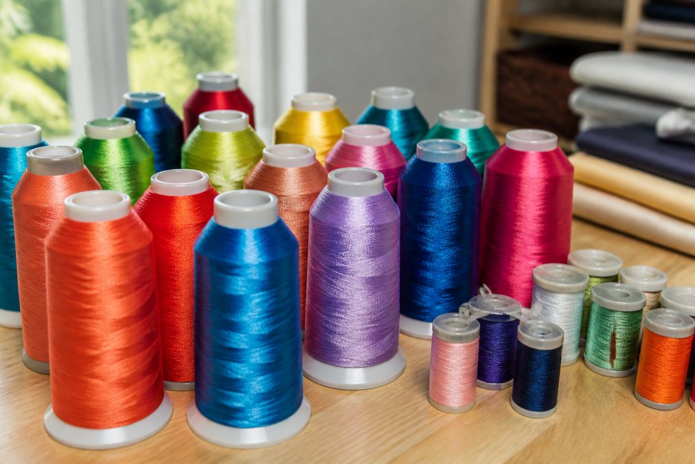 Close-up of various embroidery thread spools in multiple colors arranged on a work table, showing thread sheen and texture variations in natural daylight