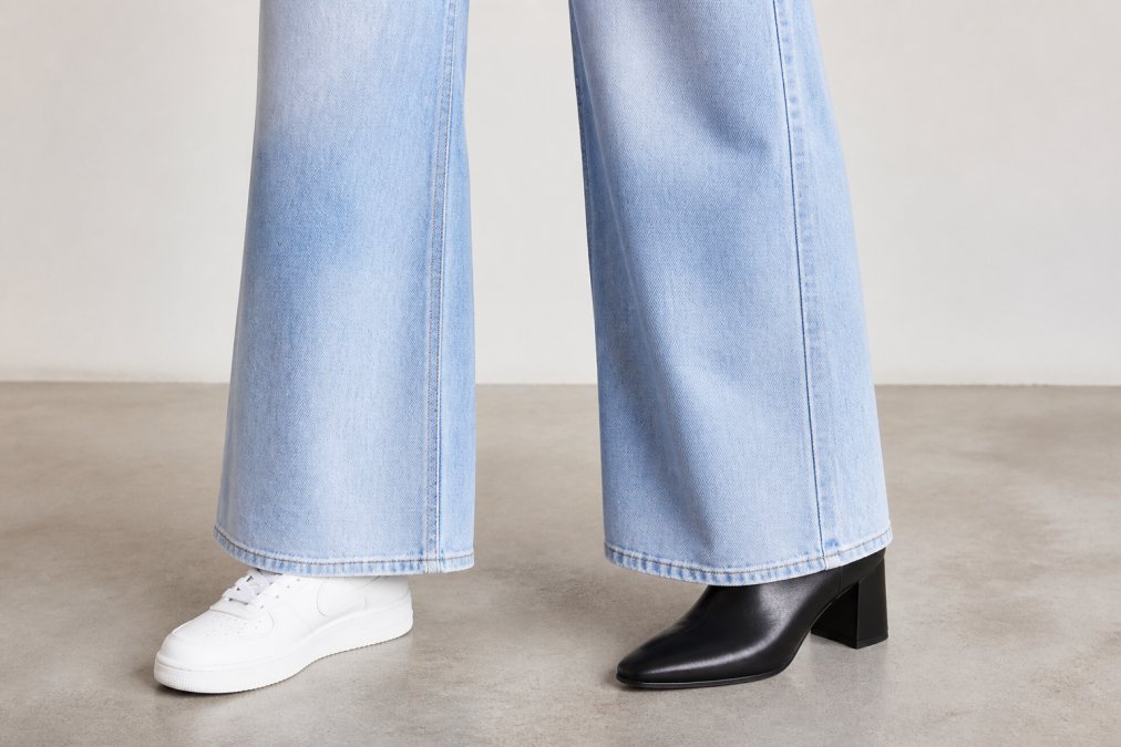 Close-up of a woman's feet showing high-waisted wide-leg jeans paired with white leather sneakers on one side and black heeled boots on the other, demonstrating contrasting shoe pairing options