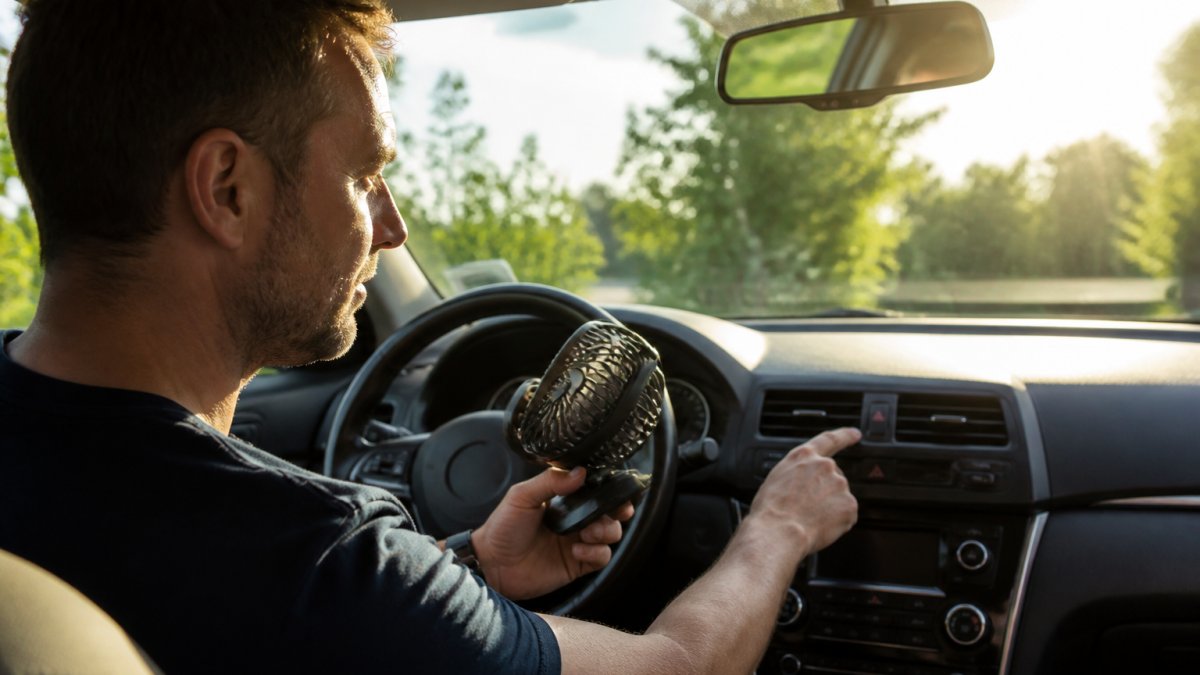 A person sitting in the driver's seat of a car, holding a small portable fan and checking a digital temperature display on the dashboard, with sunlight streaming through the windshield