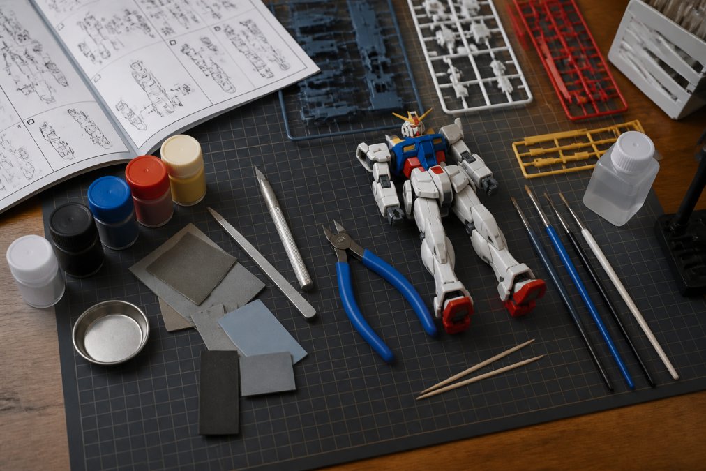 Overhead flat-lay of essential model kit tools including hobby knife, nippers, cutting mat, sanding tools, paint bottles, brushes, and a partially assembled plastic model kit on a clean workbench with reference manual nearby