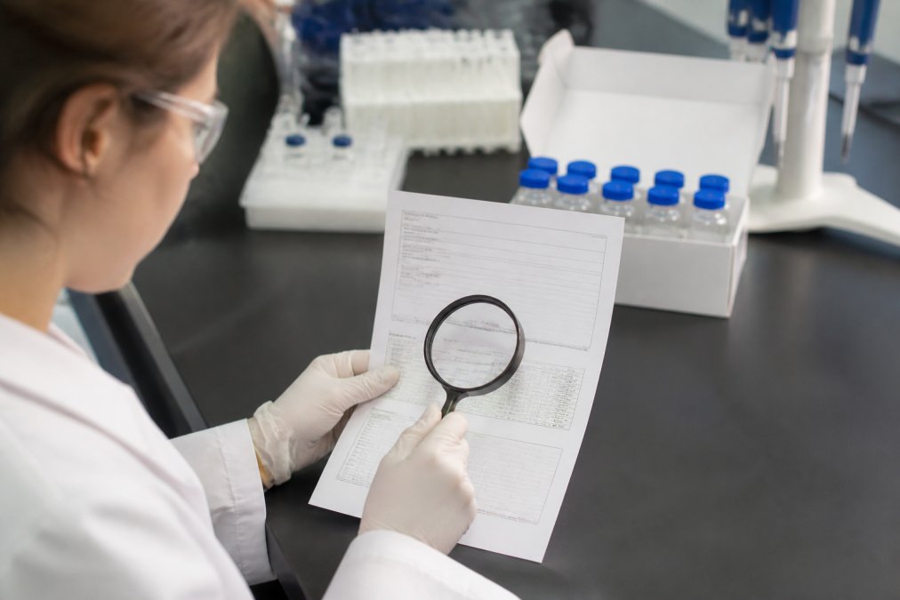Researcher examining a printed certificate of analysis document at a laboratory bench with a magnifying glass, peptide vials visible in the background