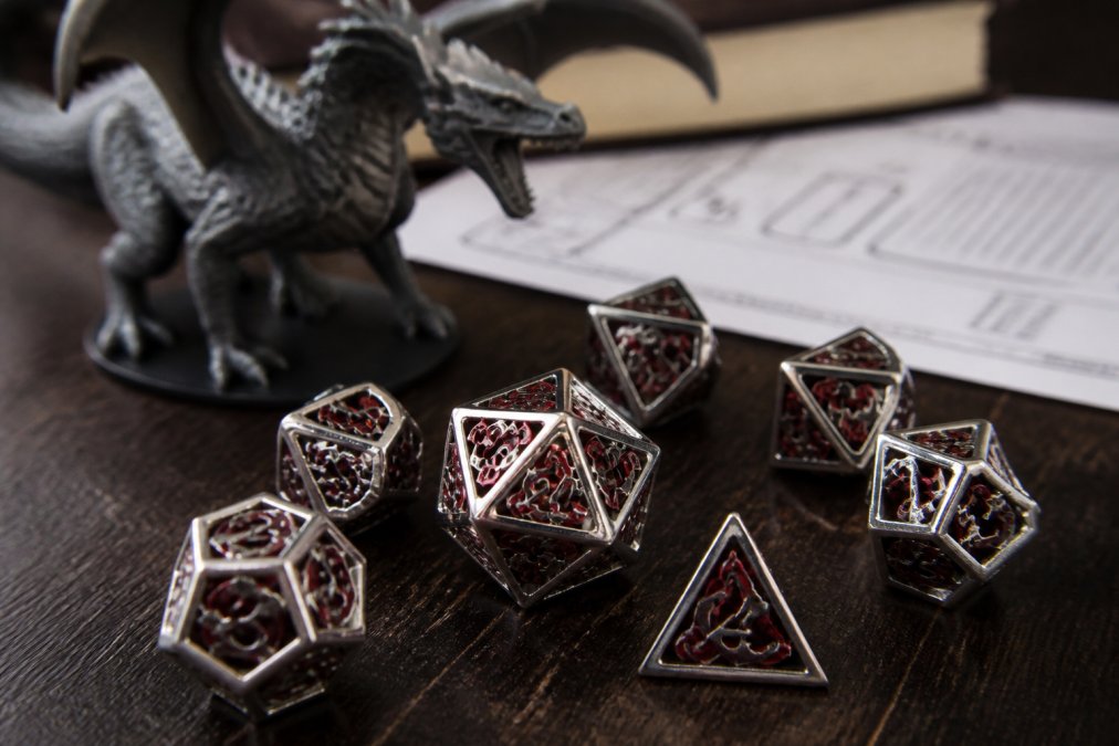 Close-up of a set of silver and red hollow metal polyhedral dice scattered on a dark wooden gaming table, with a dragon miniature and character sheet in the background