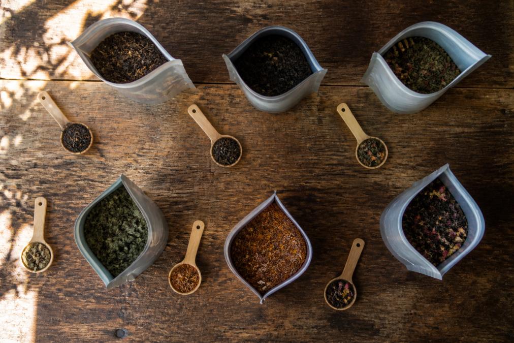 Overhead flat lay of several open resealable pouches of loose leaf tea on a rustic wooden table, with small spoons of dried tea leaves beside each pouch, natural window light casting soft shadows