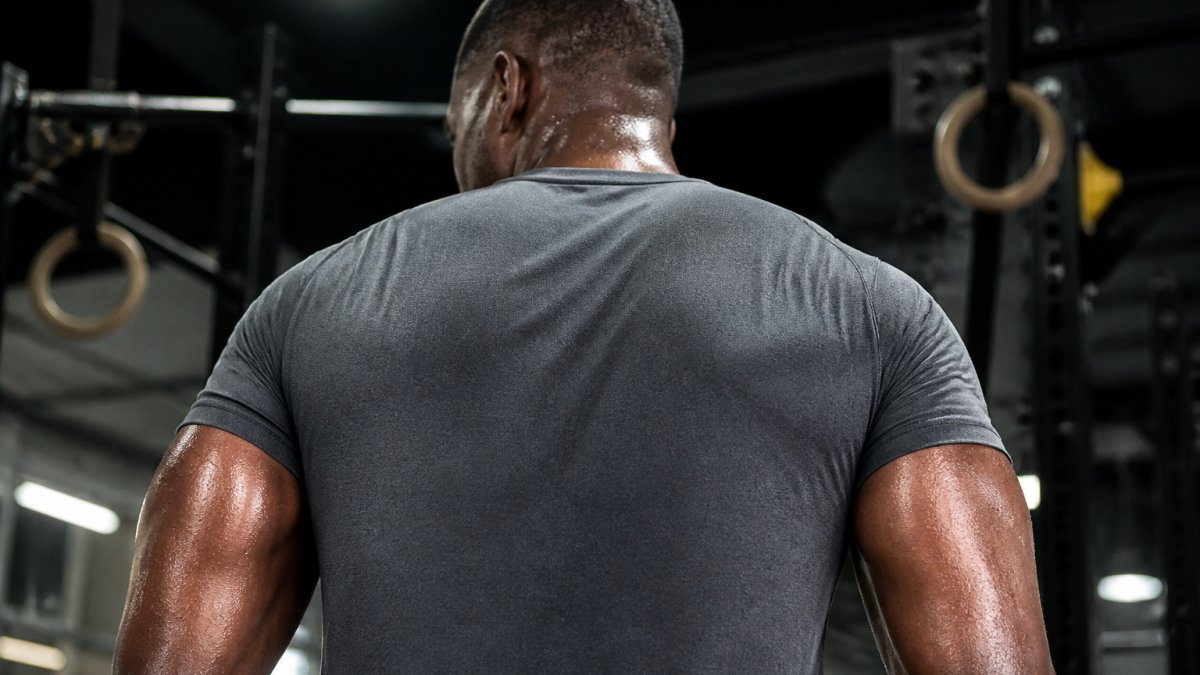 Close-up of an athlete's back and shoulders during an intense gym session, shirt visibly dry while sweat drips from skin, harsh overhead gym lighting