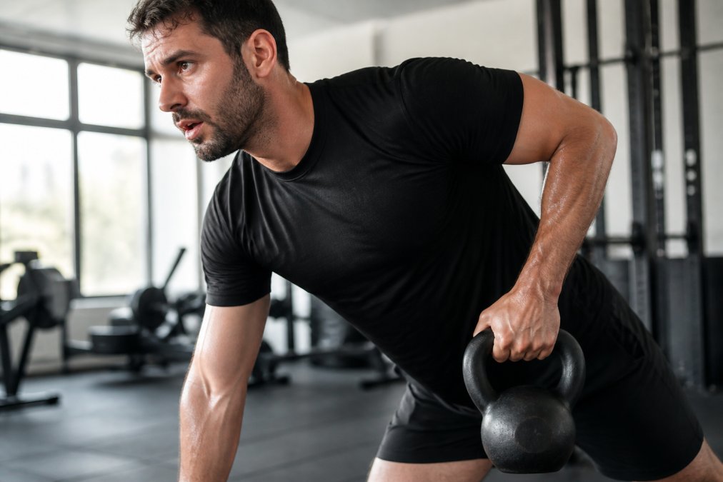 Man lifting a kettlebell in a gym wearing one of our moisture wicking gym shirts with visible sweat patterns.