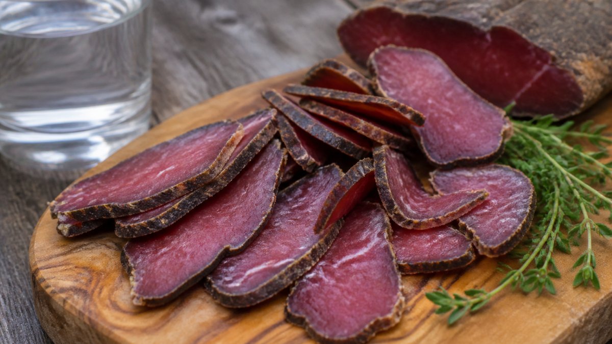 Close-up of sliced grass-fed biltong on a wooden board with fresh herbs and a glass of water, showing the deep red colour and texture of premium air-dried meat
