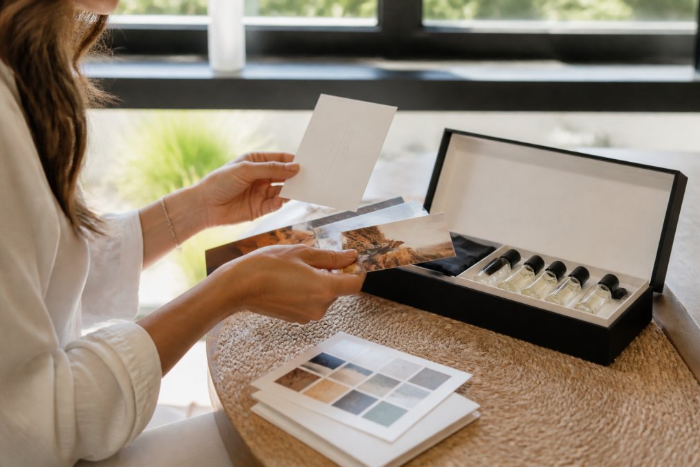 Person holding an elegant fragrance discovery set with multiple small bottles, examining narrative cards and scent notes in natural daylight near a window