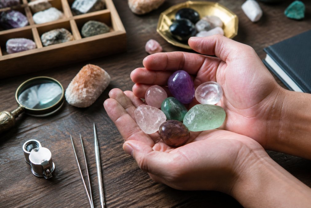 Close-up of hands holding several polished gemstones and tumbled crystals on a wooden surface, with natural light illuminating their translucency and natural formations