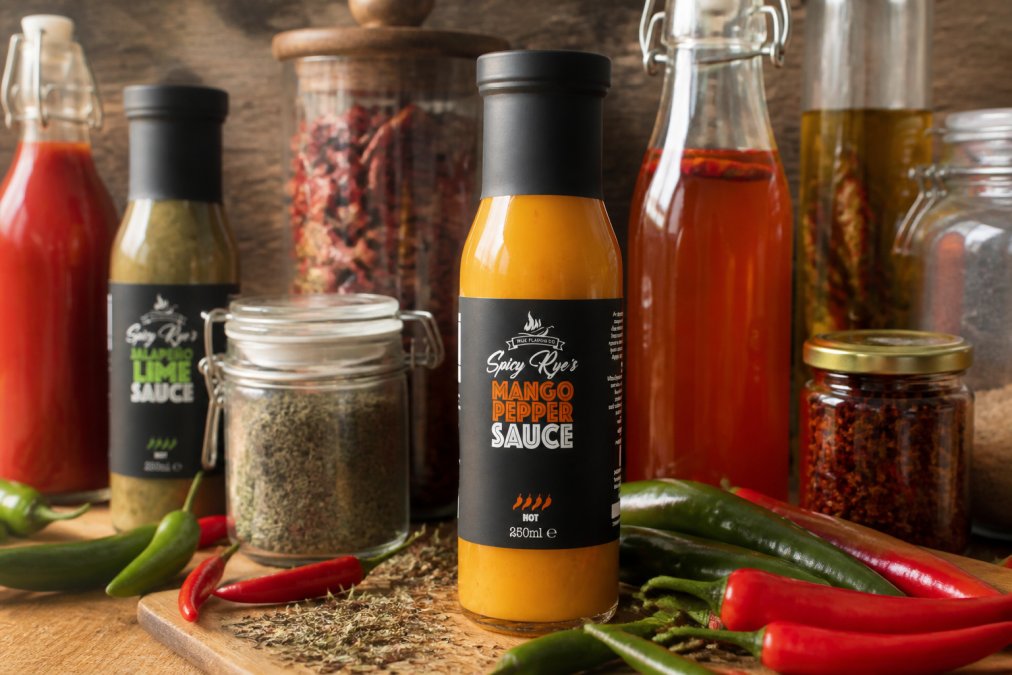 Close-up of premium spicy sauce bottles and chilli vinegars arranged on a wooden surface with fresh red and green chillies, dried herbs, and artisanal glass jars with handwritten labels catching warm afternoon light