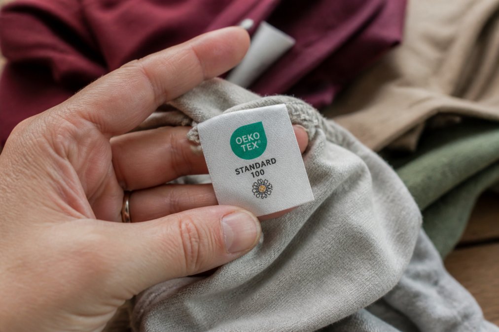 Close-up of a hand holding a fabric label with the OEKO-TEX Standard 100 logo, soft natural light, casual cotton clothing blurred in the background