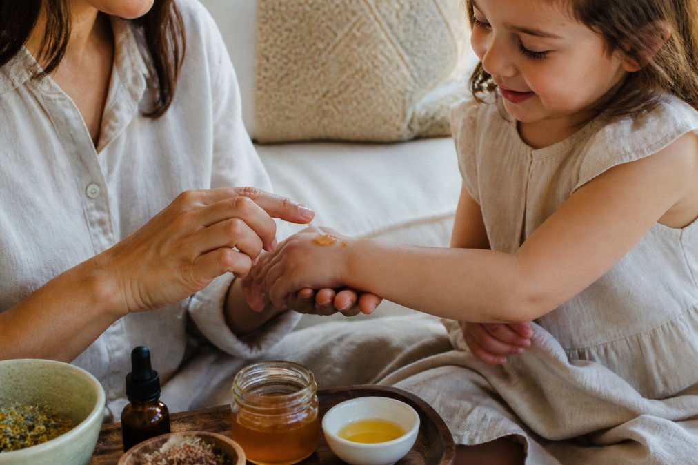 Close-up of a parent's hands gently applying a small amount of honey-based balm to a young child's forearm, soft natural light, cozy home setting, no visible skin condition