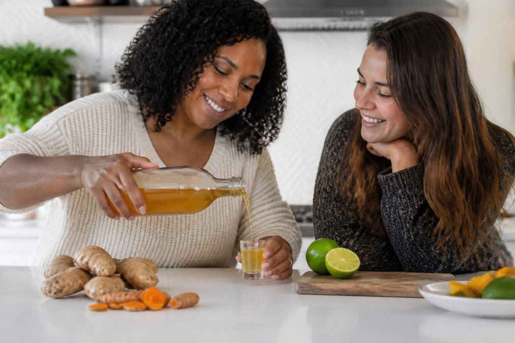 A hand pouring golden amber tonic from a glass bottle into a small shot glass on a bright kitchen counter, fresh ginger root, turmeric pieces, and a halved lime nearby in soft natural light