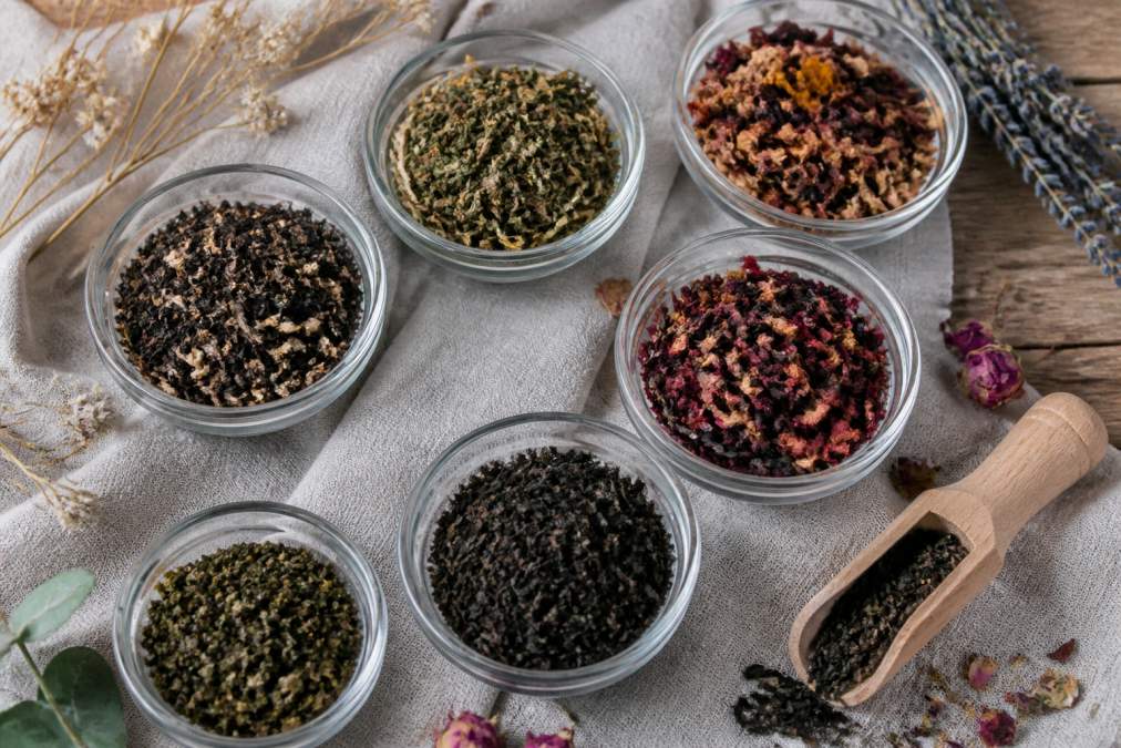 Overhead flat-lay of several small glass bowls filled with different organic loose leaf teas, green, black, oolong, and herbal blends, arranged on a natural linen cloth with a wooden scoop and dried botanicals