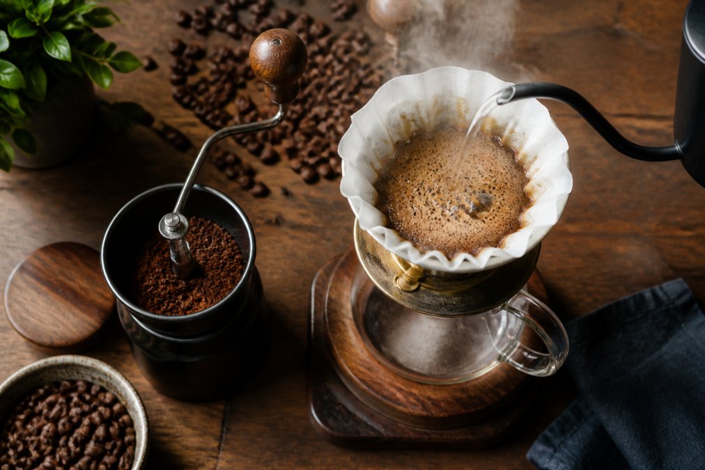 Overhead view of a pour-over cone with whole coffee beans scattered nearby, a burr grinder holding freshly ground beans, and hot water being poured in a thin stream creating steam and foam
