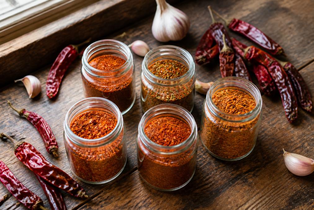 Overhead flat-lay of several small glass spice jars filled with vibrant red and orange seasoning blends arranged on a rustic wooden kitchen counter, with dried red chili peppers and whole garlic cloves scattered around them, warm natural light from a nearby window