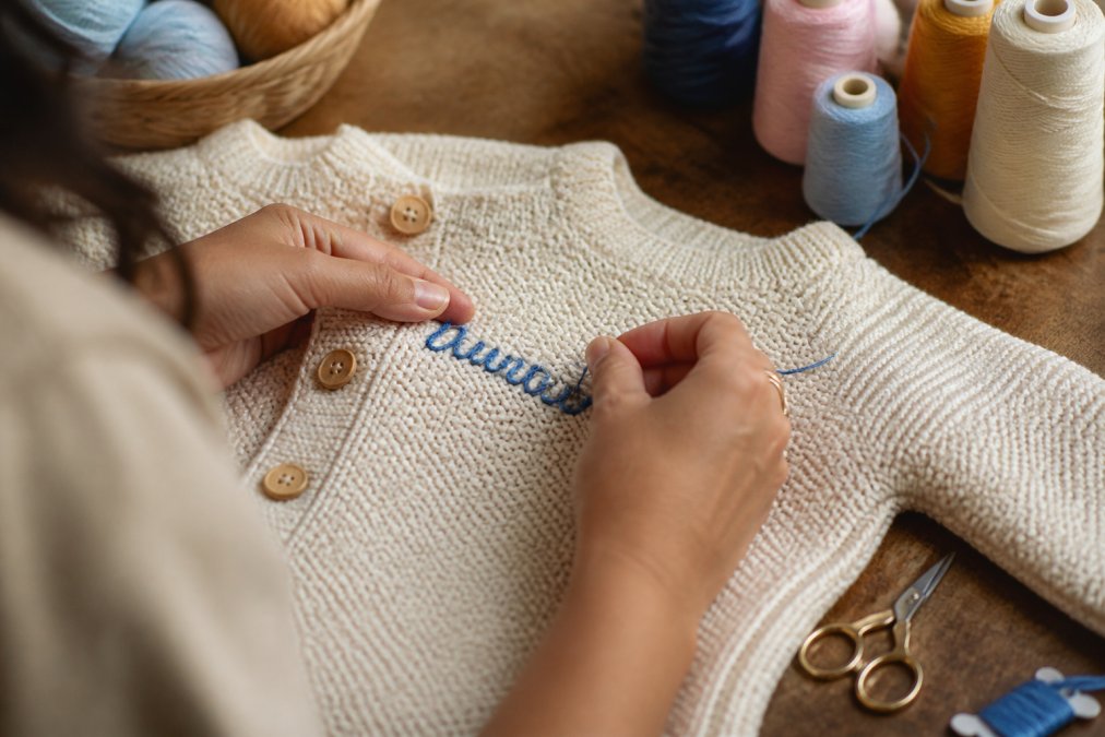 Close-up of a craftsperson's hands carefully embroidering a baby's name in blue thread onto a cream-colored hand-knitted sweater, colorful yarn spools and small embroidery scissors arranged on a warm wooden worktable beside the piece