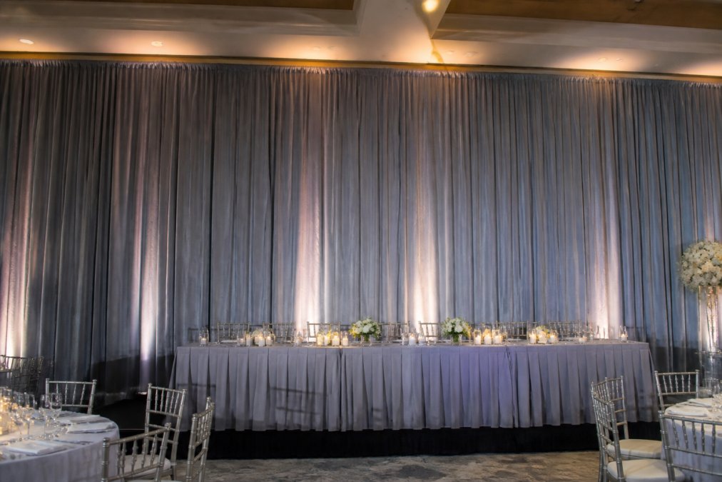 Professional event backdrop with elegant velour drapes in neutral gray displayed at a wedding reception, with soft ambient lighting and decorative uplighting illuminating the fabric