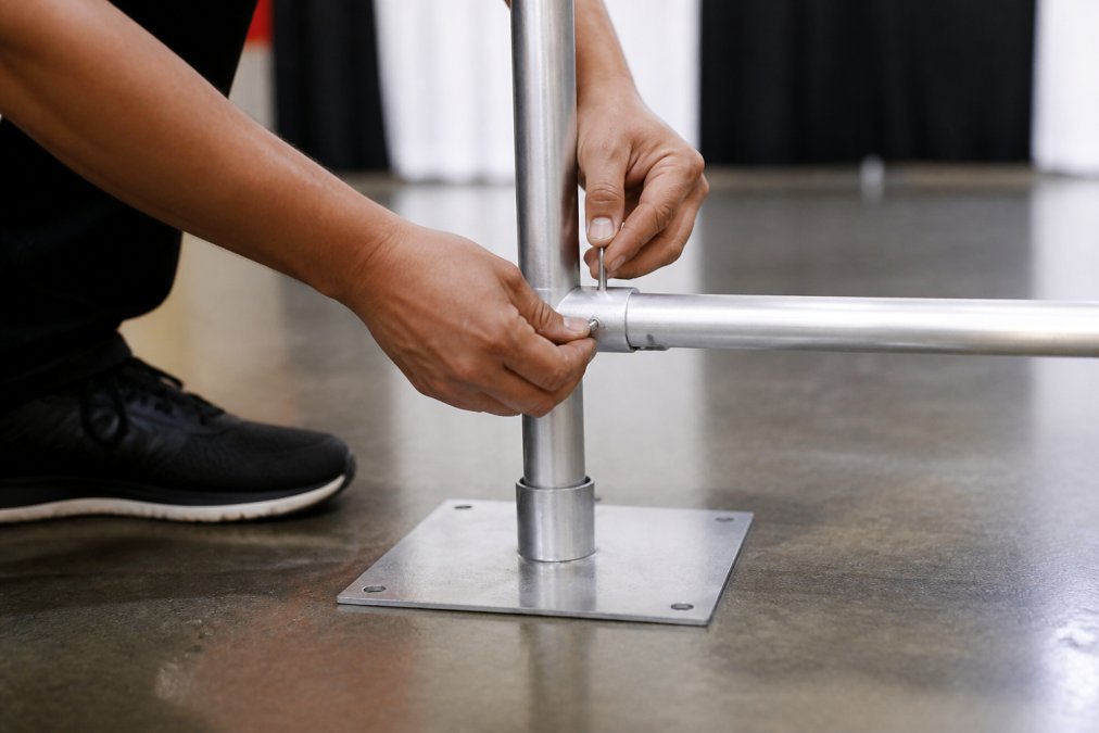 Close-up of hands carefully assembling the base plate, upright pole, and crossbar connection of a pipe and drape system, showing the button-lock mechanism engagement during professional setup