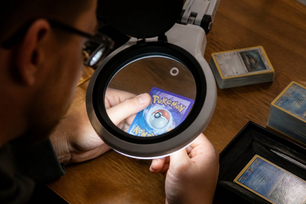 Professional examining a Pokemon trading card under bright magnifying light, comparing card texture and printing details on a wooden table