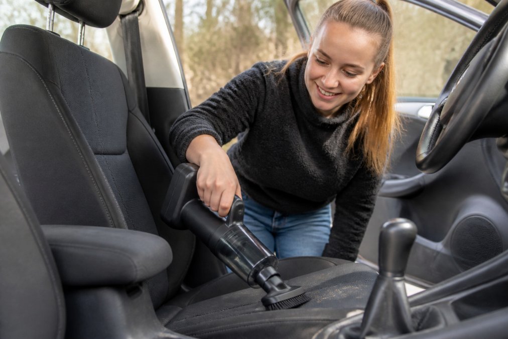 A person holding a compact cordless handheld vacuum inside a car, cleaning the seat upholstery with a brush attachment, natural daylight coming through the car window
