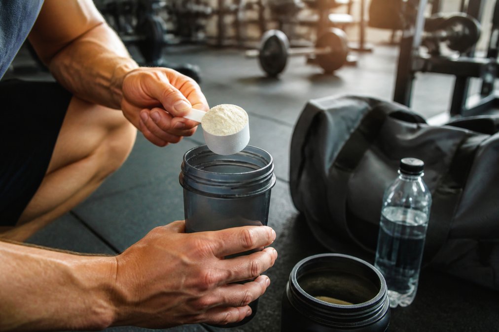 A gym-goer's hands holding a shaker cup with pre-workout powder and a scoop, with a training bag and water bottle in the background