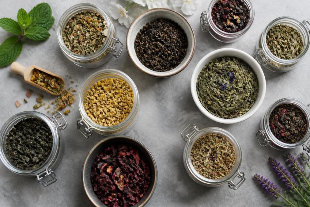 Overhead flat lay of various loose leaf tea types in clear glass jars and ceramic containers, showing different colors and textures of premium tea leaves with fresh herbs and flowers scattered nearby