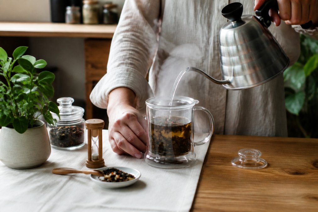 Close-up of hands pouring hot water from a kettle into a glass infuser filled with loose leaf tea, steam rising, with a timer and brewing guide visible nearby