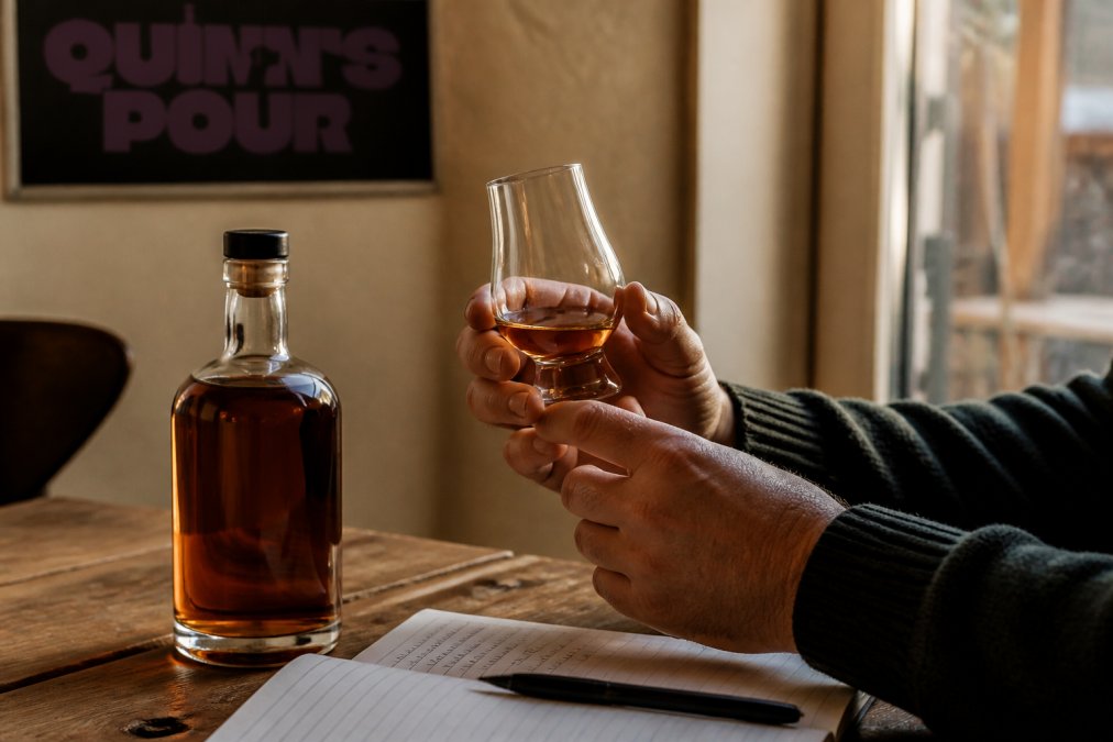 Close-up of a person's hands holding a glass of spiced rum, with a bottle of Quinn's Pour rum and a tasting notebook on a wooden table in a warm, softly lit room