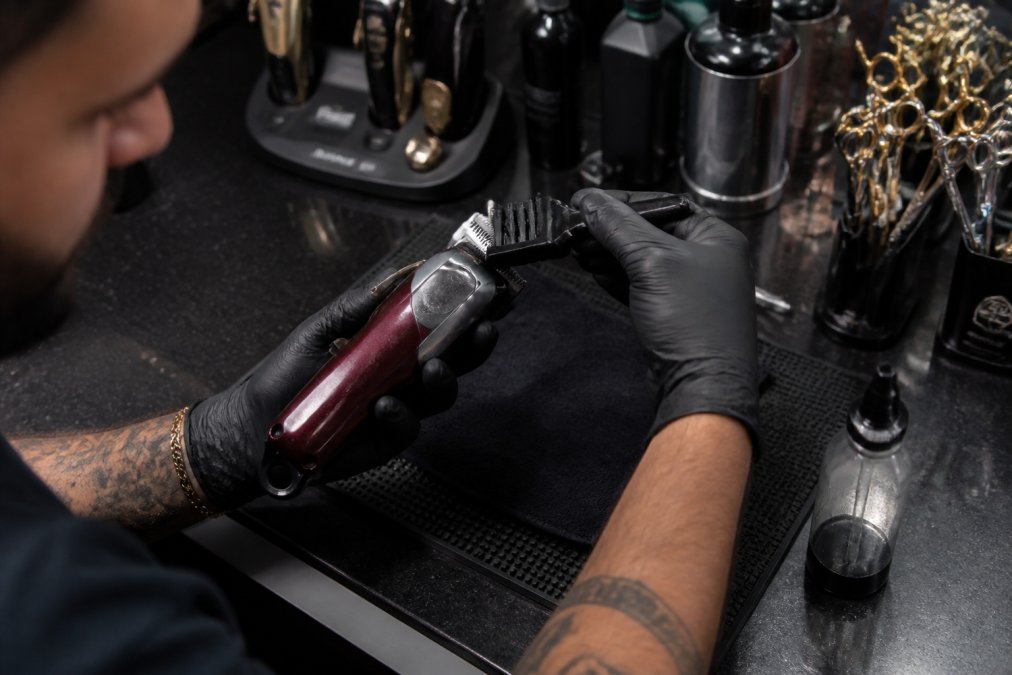 Barber carefully cleaning and maintaining clipper blades with oil and brush on a clean station counter, with organized grooming tools and supplies visible nearby