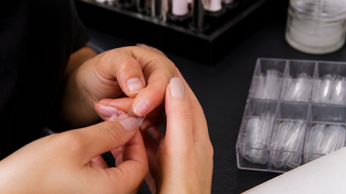 Close-up of a nail technician's hands holding a soft gel nail tip against a client's natural nail to check fit, with a tidy salon workstation and tip boxes blurred in the background