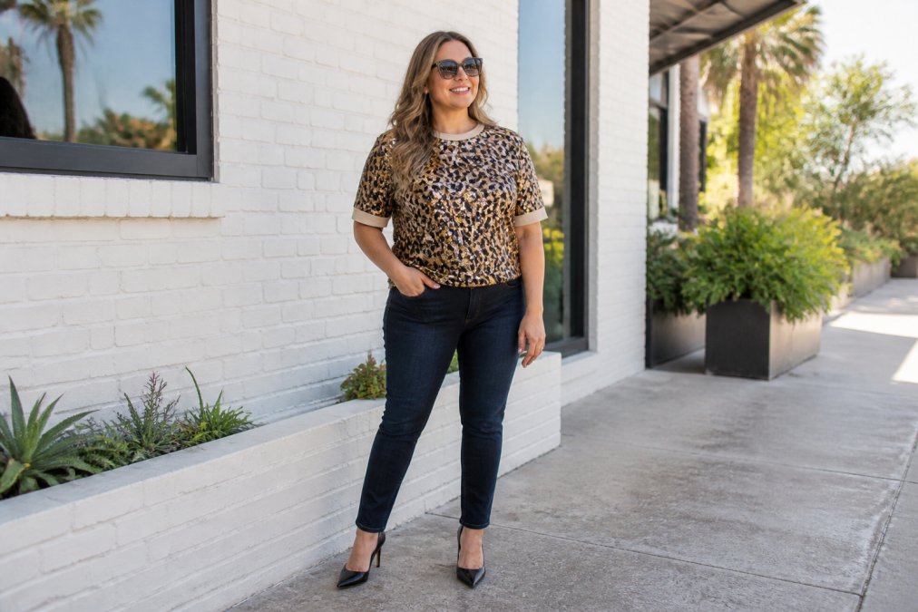 Woman wearing a Queen of Sparkles sequin leopard tee styled with dark fitted jeans and black leather heels, standing in natural daylight with confident posture