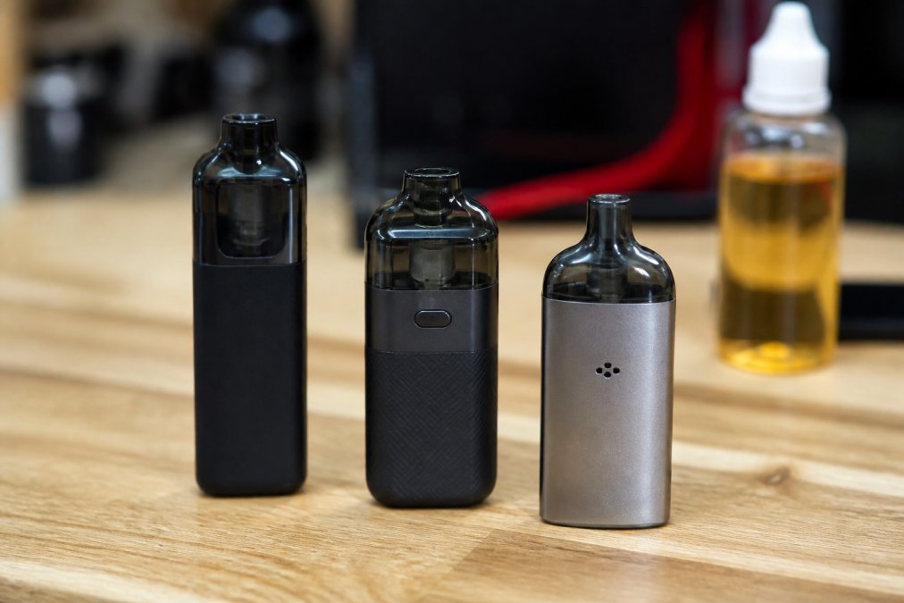 Close-up of three compact refillable pod vape devices arranged on a light wooden table, showing different sizes and finishes, with a bottle of e-liquid softly blurred in the background