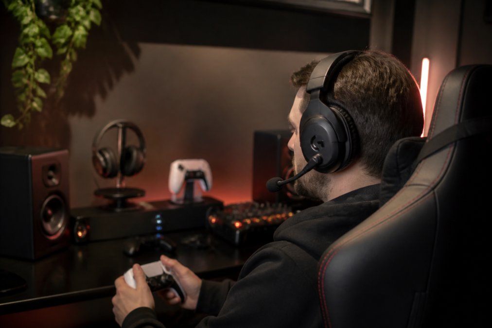 A gamer wearing a wireless gaming headset while seated at a gaming setup with multiple monitors, RGB lighting, and gaming peripherals visible in the background under warm studio lighting