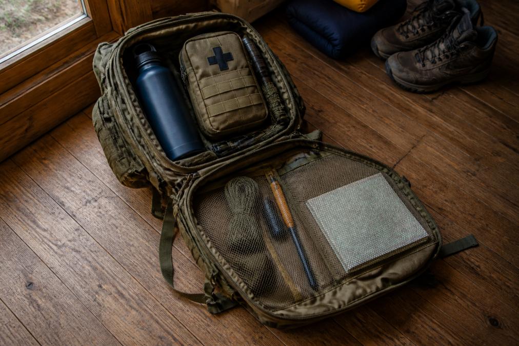 Overhead view of a fully packed tactical backpack opened on a wooden cabin floor, showing organized compartments with a water bottle, first aid pouch, ferrocerium rod, and folded map, lit by warm natural window light