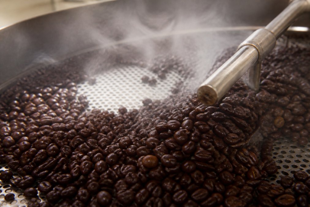 Close-up of freshly roasted coffee beans cooling on a perforated metal tray, steam rising from the beans, showing deep mahogany and chocolate brown colors with glossy surfaces reflecting light