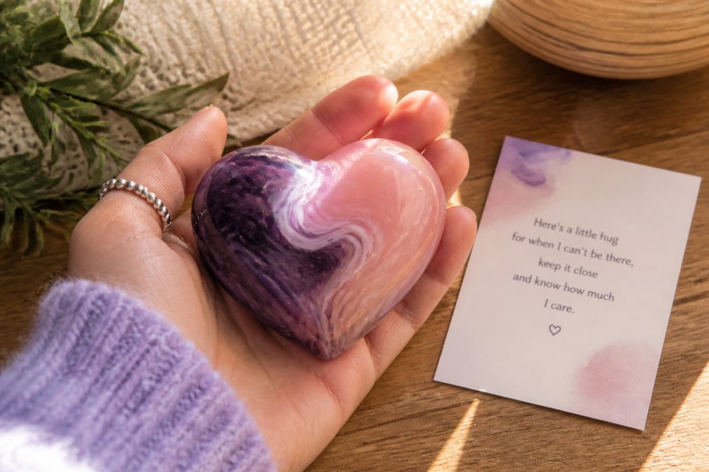 Close-up of a hand-finished heart-shaped resin keepsake in soft blush and cosmic purple colours, resting in an open palm with natural morning light highlighting the swirled texture, accompanied by a small poem card placed beside it