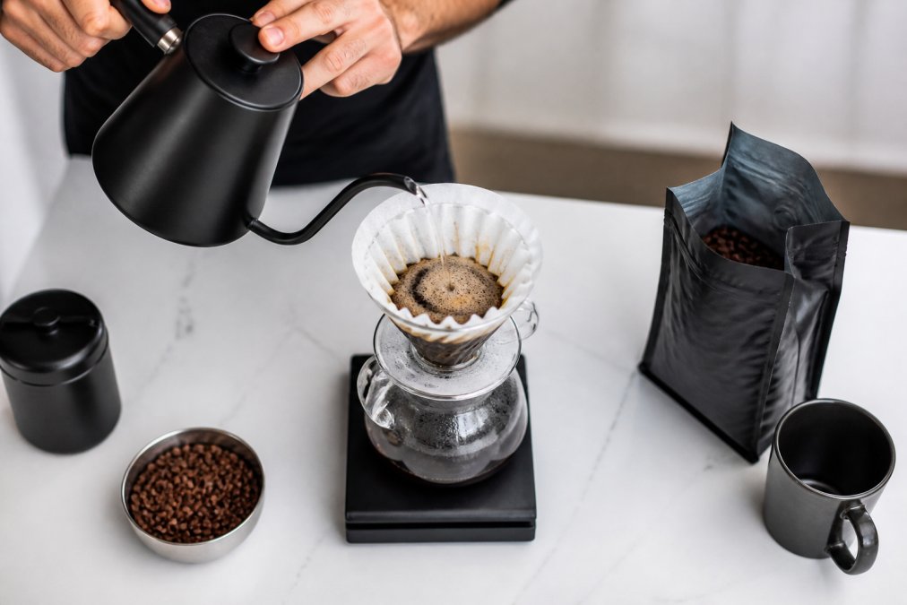 Overhead shot of hands pouring hot water from a gooseneck kettle through a pour over dripper set on a glass carafe, surrounded by whole bean coffee and a small digital scale on a clean white countertop
