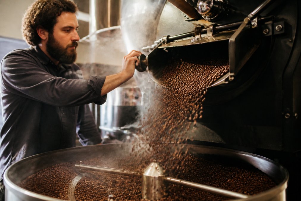 Coffee roaster pouring freshly roasted beans into a cooling tray with steam rising, highlighting the roasting process and the moment beans transition from dark brown to their final color