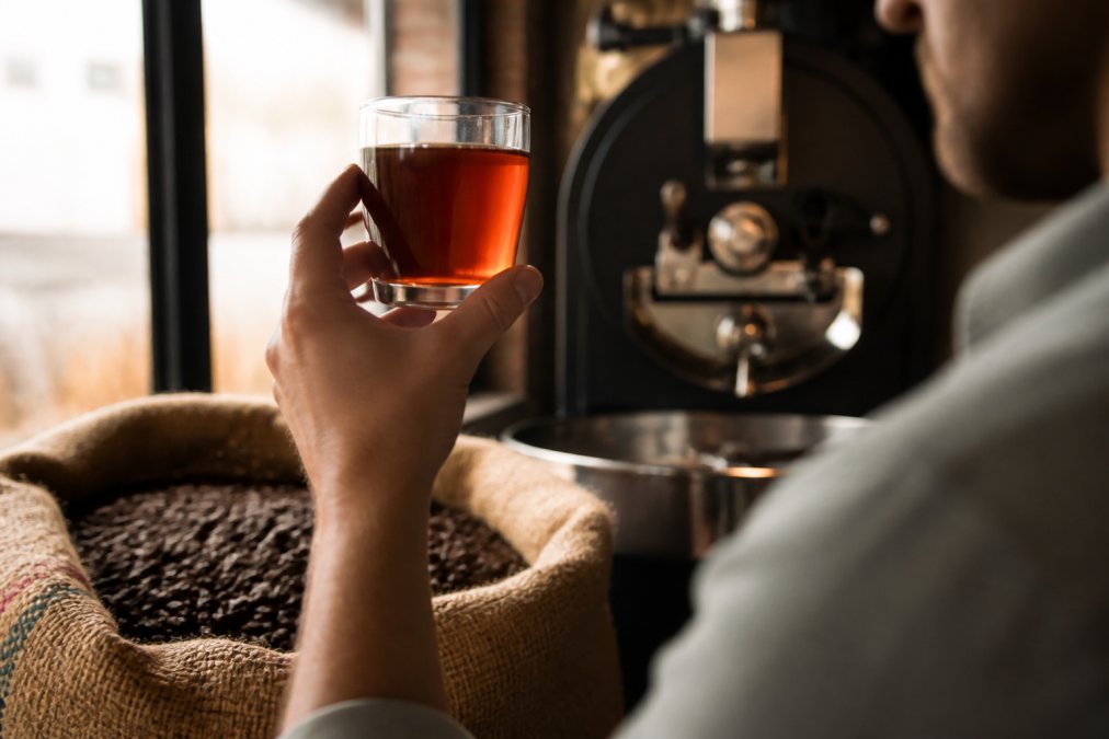 A person holding a clear glass of freshly brewed coffee up to the light, with a burlap sack of coffee beans and a roasting machine softly blurred in the background